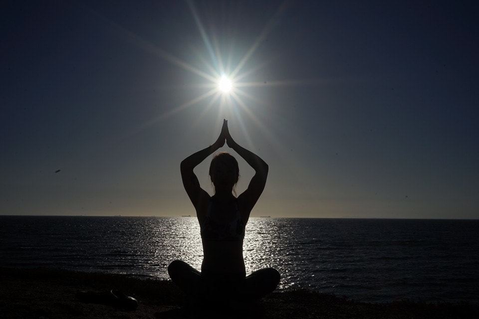 A figure meditating in silhouette by the sea, hands joined above the head, with the sun low in the sky